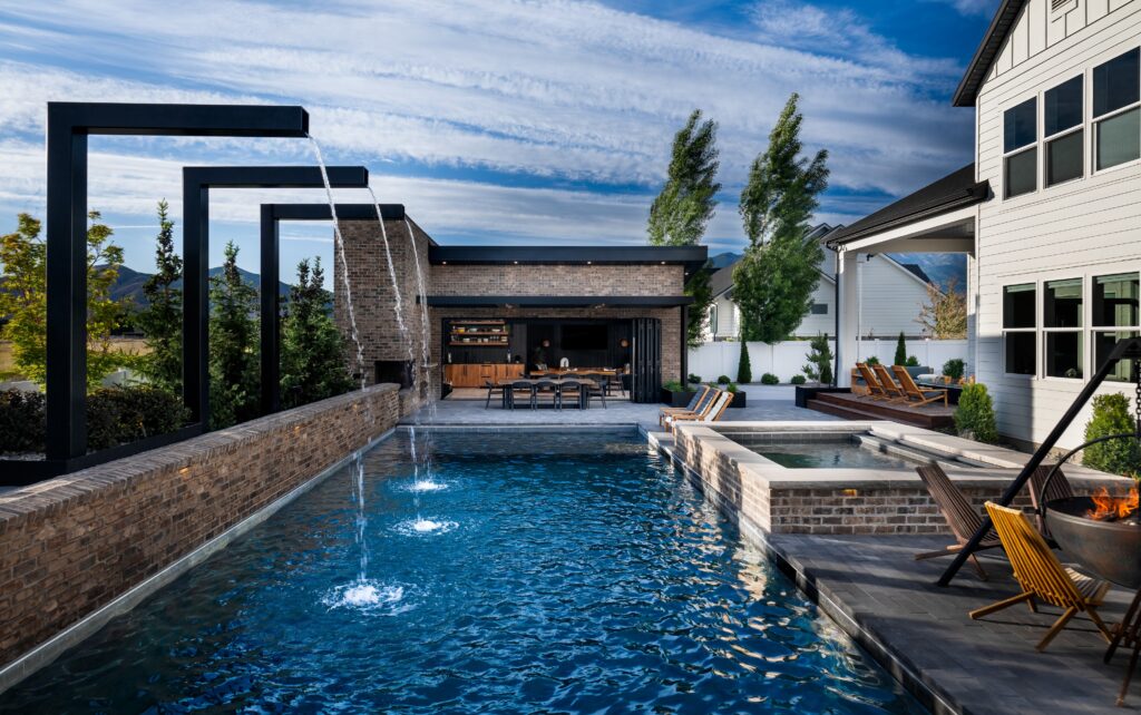 A backyard with luxurious pool with modern fountains, an outdoor kitchen, hot tub, and accent trees