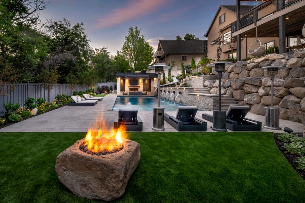 A backyard with fire pit contained in a large rock, pool with cascading fountains and tiered landscaping with large rock accents