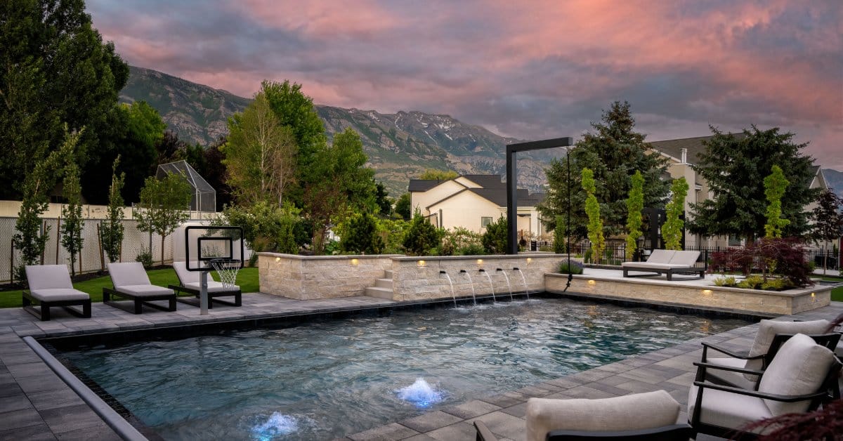 A sunset view of an in-ground pool. The sky is orange and purple and streaky and the pool has water features on the edge.