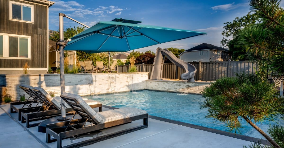 A small backyard pool with a slide in the corner and a large canopy umbrella on the patio area with three chaise lounges.