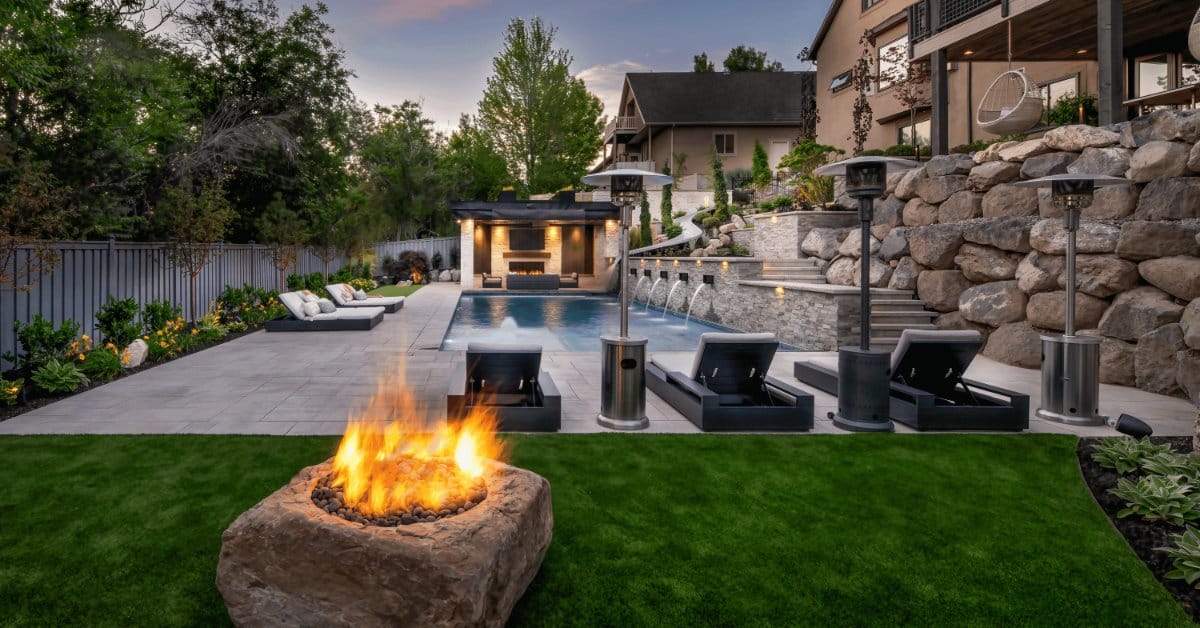 A small side yard next to a luxury in-ground pool. There's a stone fire pit burning and three chaise lounges by the pool.