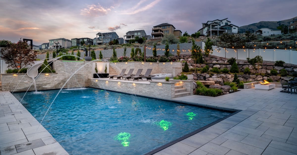 A nighttime view of the backyard with a luxury pool lit up and patio lights strung up. A water feature shoots into the pool.