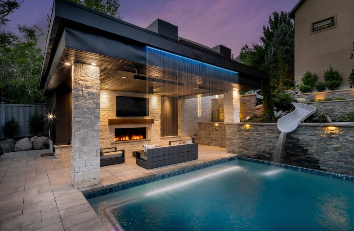 A waterfall flows from the top of an outdoor pavilion and flows into the pool below. A slide also flows into the pool.