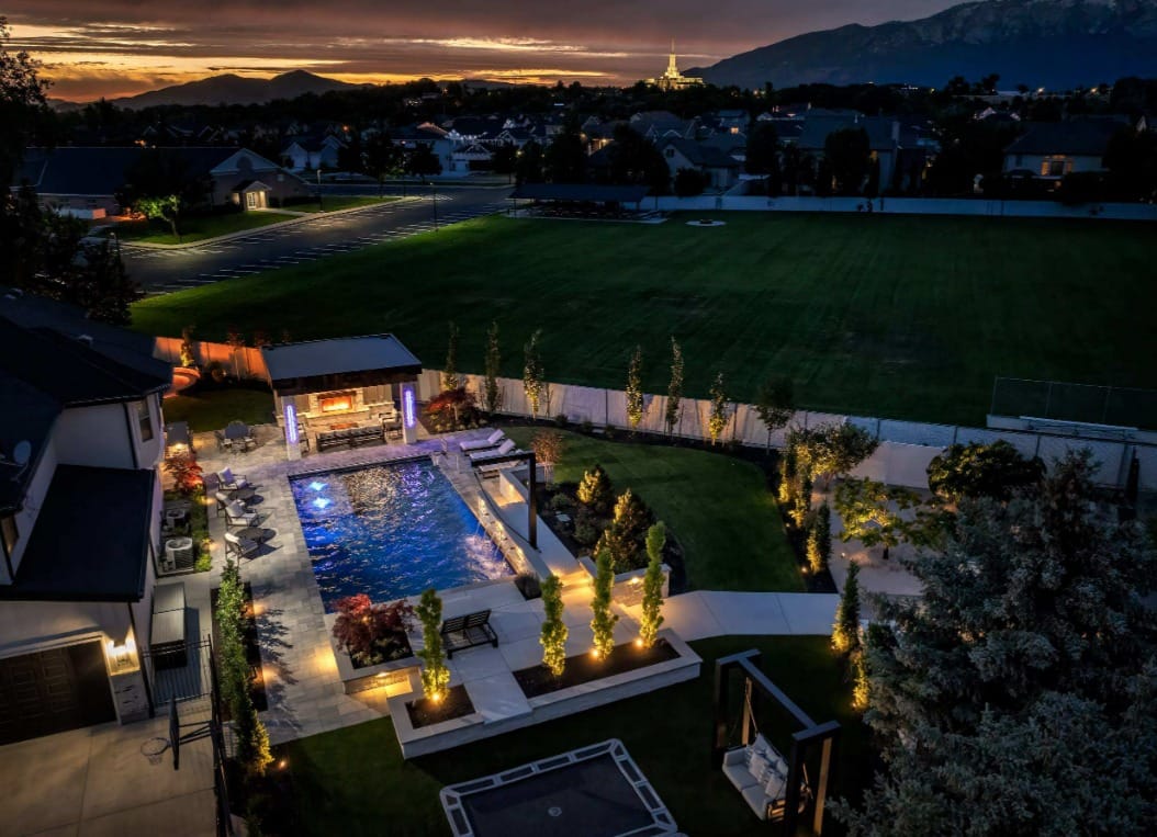 A nighttime aerial view shows a beautiful backyard with an in-ground pool, a pavilion, a fence, and beautiful trees.
