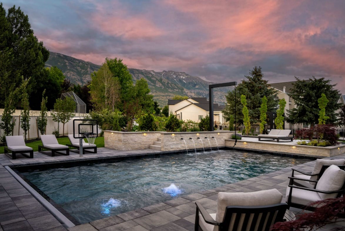 An in-ground pool features several fountains and gray tile around it. The yard is beautifully landscaped and fenced.