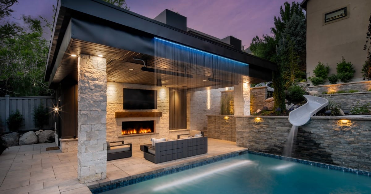 A luxury patio with a custom pergola. The pergola features a fireplace, TV, and seating, next to the pool and water feature.
