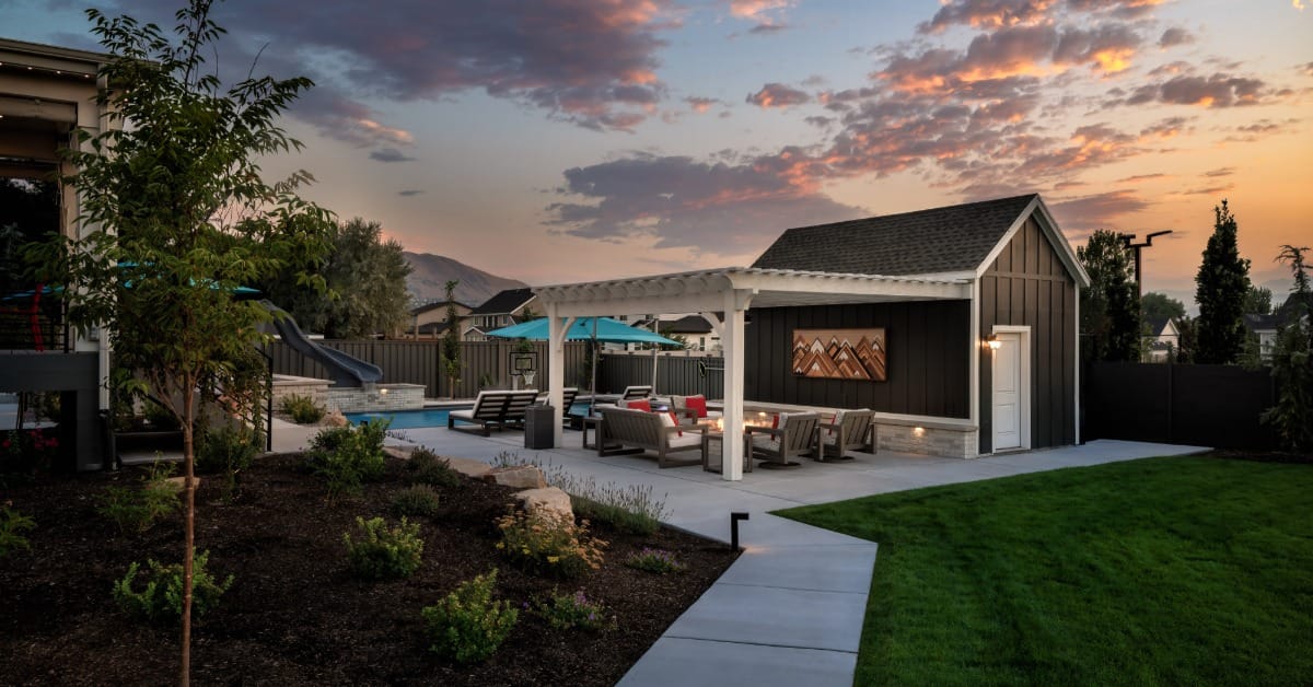 A view of the backyard sidewalk that leads to the pergola. The pergola contains comfortable seating and a shed.