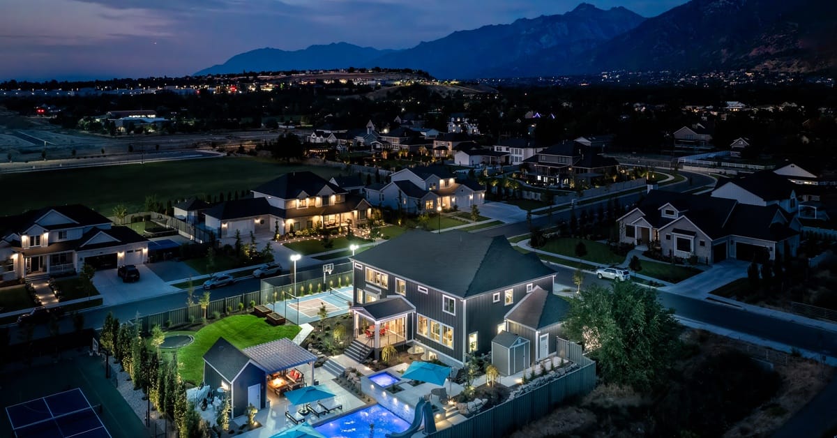 An aerial view of a luxury home in Utah. The sky is dark and the backyard features lights and a large pool with a slide.