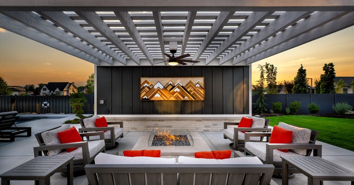 A view of an outdoor fireplace area under a pergola. The furniture matches with white cushions and red pillows.