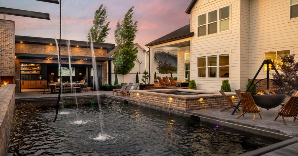 A rectangle luxury pool in the backyard of a two-story white house. There are waterfalls dropping into the pool.