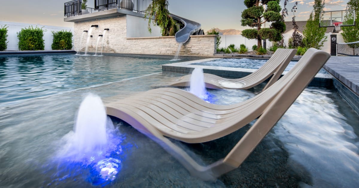 A backyard pool with two submerged lounge chairs, blue-lit fountains, a slide, stone walls, and a spa area.