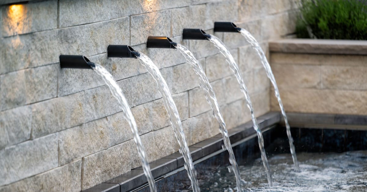 Five black spouts pouring water from a stone wall into a pool, with warm lights and greenery nearby.