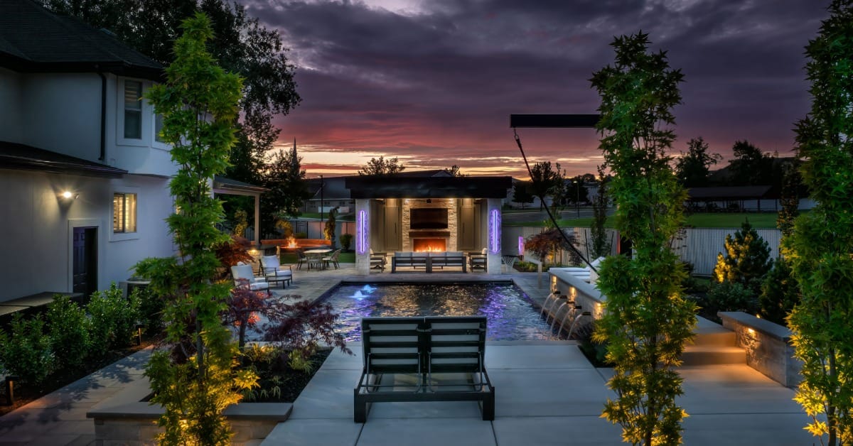 A landscaped backyard pool at dusk with lit trees, lounge chairs, a fire feature, and water spilling from a wall.