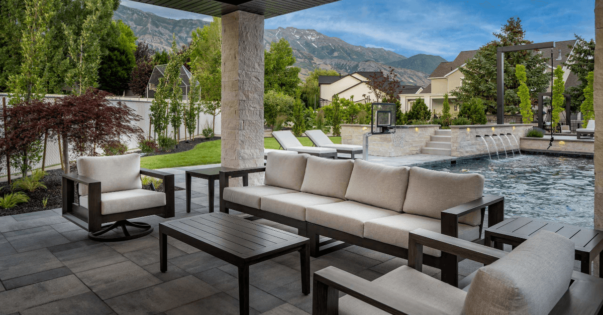 Outdoor seating under a permanent pergola in front of a luxury pool in the backyard. There are houses behind the pool.