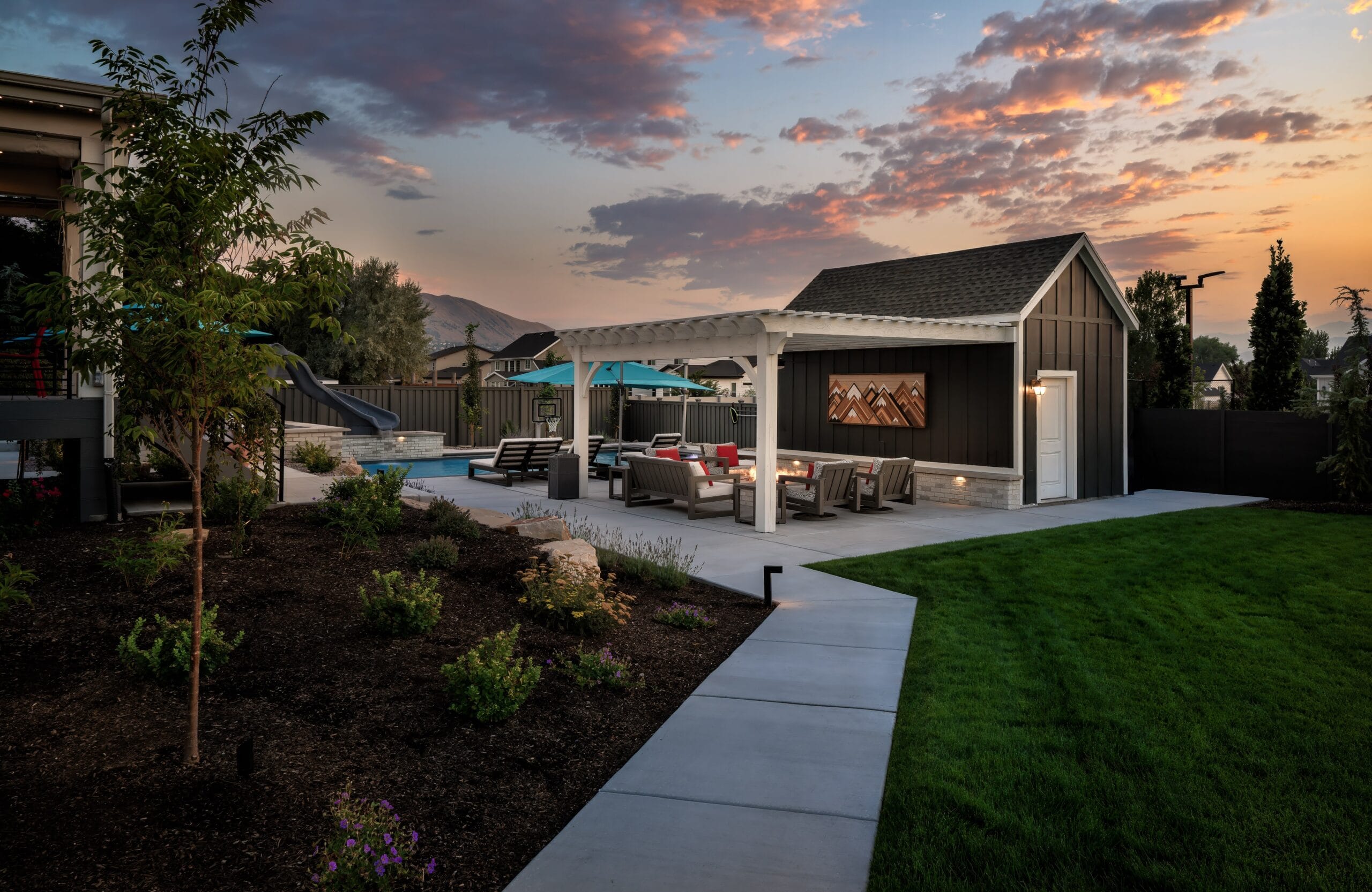 Landscaped backyard with a swimming pool, covered outdoor lounge and fire pit, green lawn, and mountain views beneath a colorful sunset.