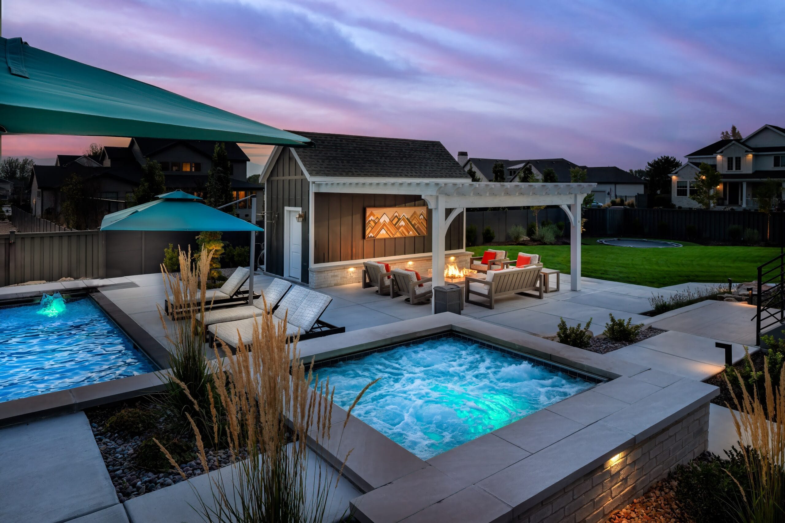 Modern backyard retreat with an illuminated spa and pool, lounge chairs, pergola-covered fire pit seating, and landscaped lawn at sunset.