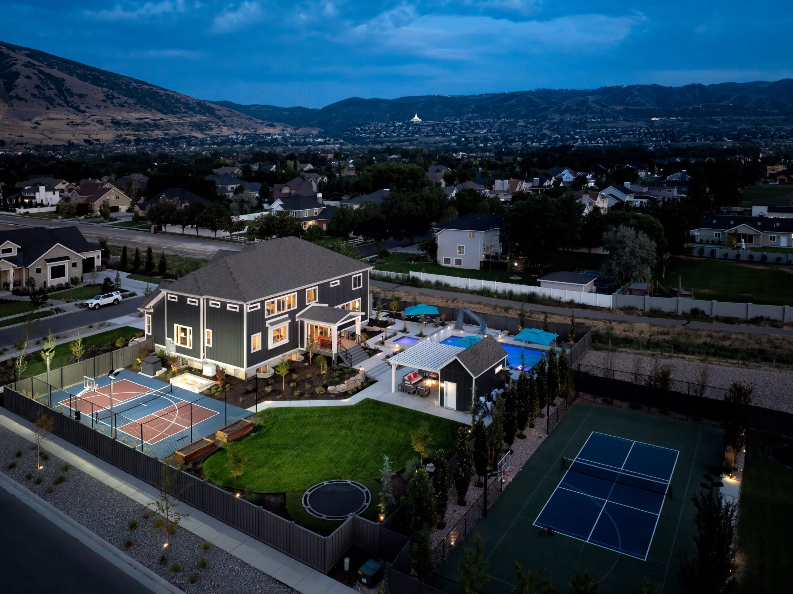 Aerial view of a luxury backyard with a swimming pool, sports courts, lawn, patio, and outdoor living areas surrounded by mountain views.