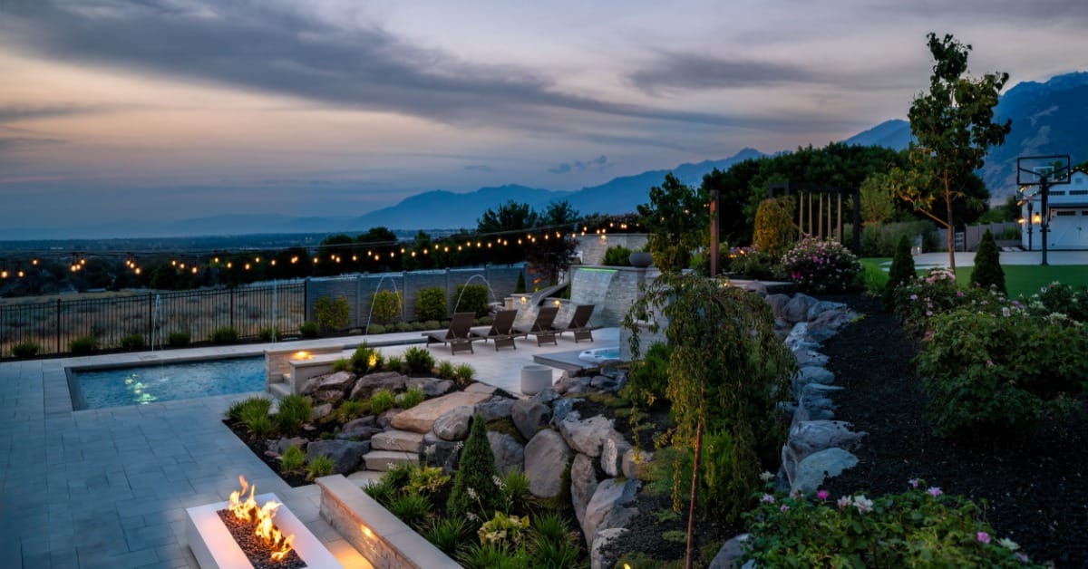 Luxury backyard pool and spa at dusk with a fire feature, lounge seating, string lights, landscaped gardens, and mountain views.