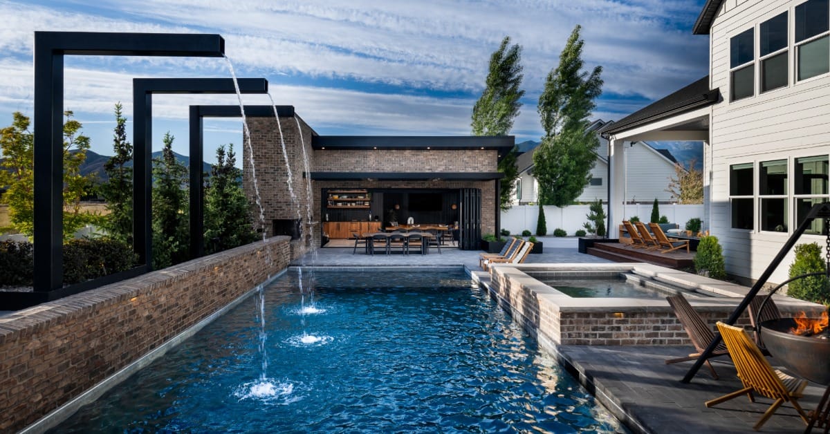 Modern backyard swimming pool with cascading water features, raised spa, outdoor kitchen, lounge seating, and fire pit.